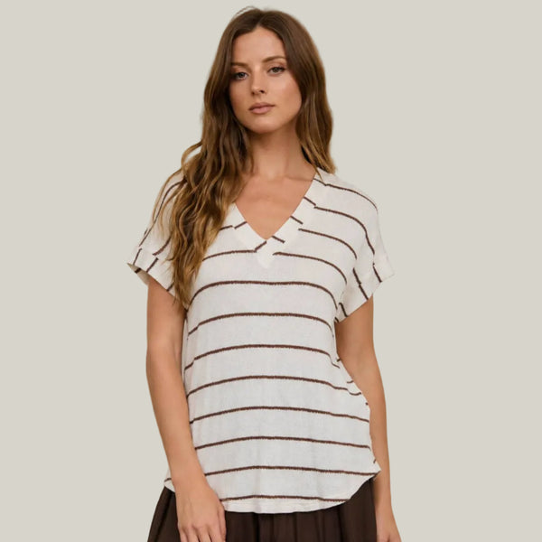 A woman wearing a white short-sleeve V-neck top with thin brown horizontal stripes, paired with dark brown wide-leg pants, standing against a beige background