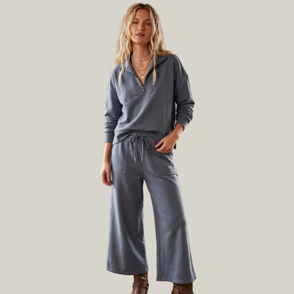 A woman stands wearing a matching dusty blue-gray lounge set consisting of a long-sleeve drawstring henley top and wide-leg drawstring pants, paired with brown leather boots