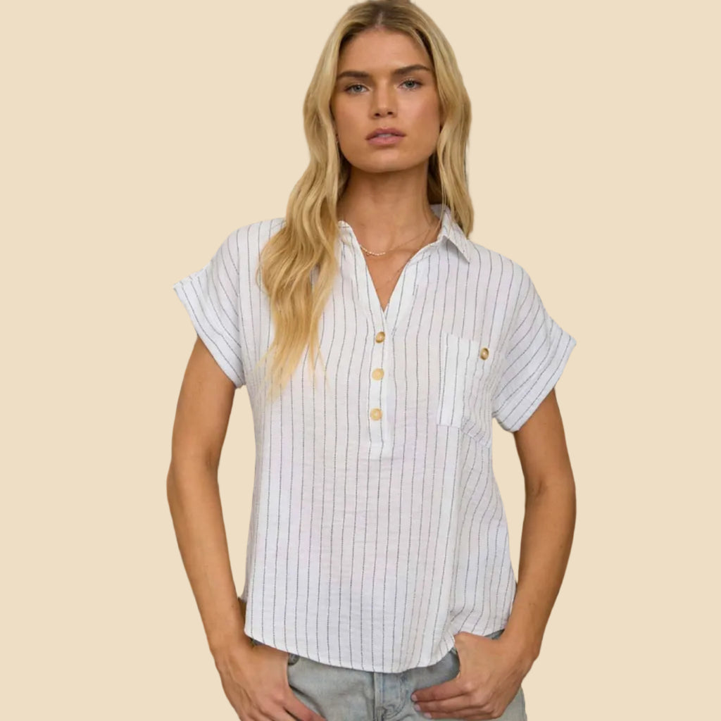 A blonde woman stands indoors wearing a white short-sleeve button-front shirt with thin vertical dark pinstripes and light denim shorts. 