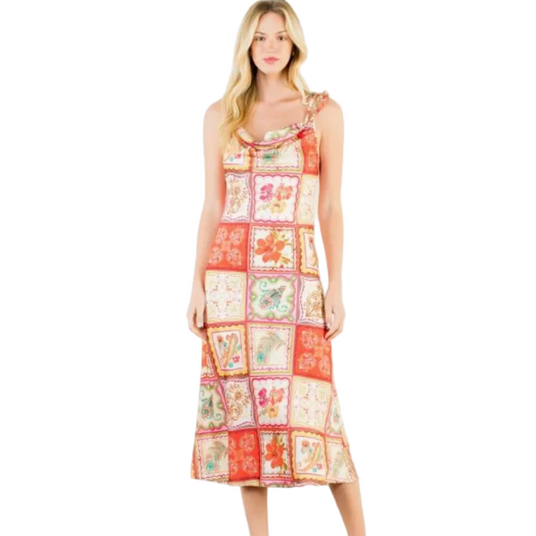 A woman modeling a sleeveless midi dress with a colorful patchwork print of floral and geometric tiles in orange, red, pink, and cream, set against a plain white studio background.
