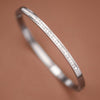 A polished silver-colored metal bangle bracelet with a row of small square-cut stones set along the outer band, displayed on a warm brown background.