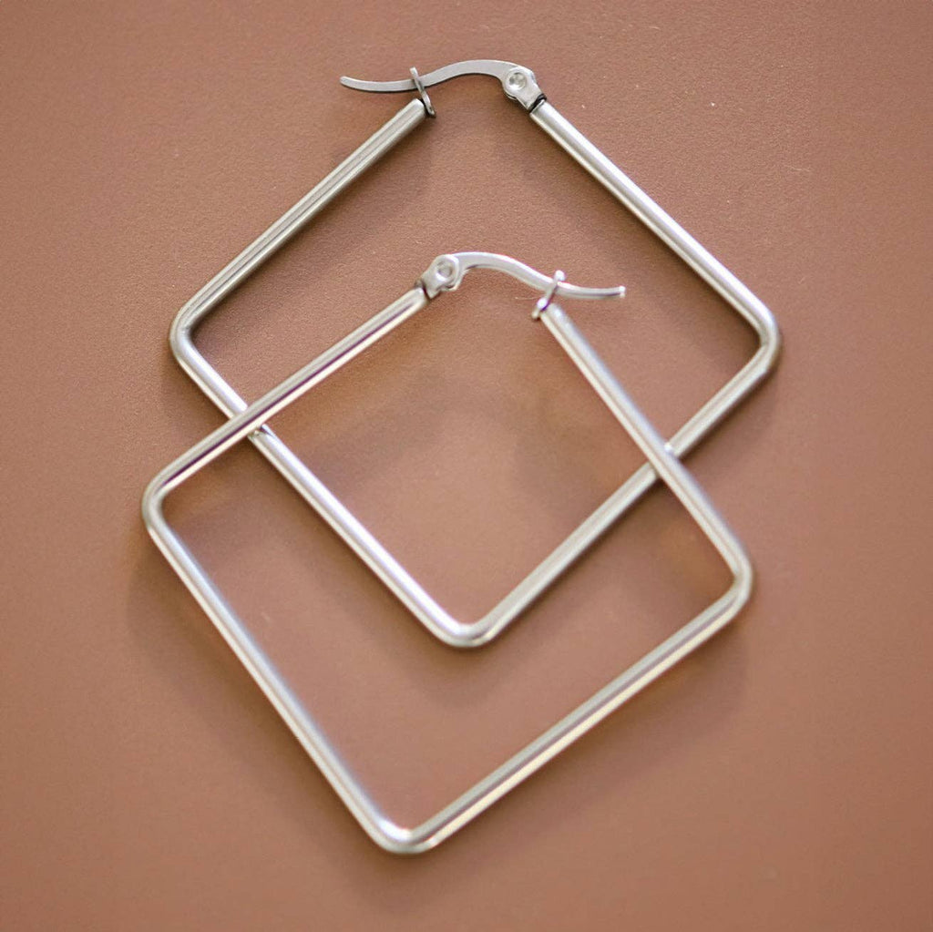 A pair of silver-toned geometric hoop earrings with a double-line square design, displayed on a solid warm brown background.
