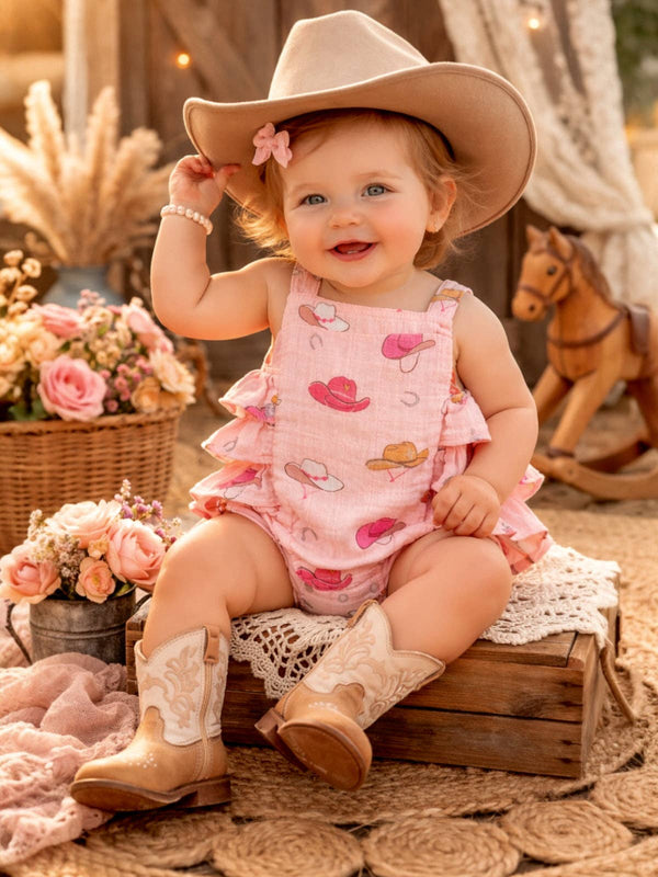 Baby in pink romper and cowboy hat sitting on a wooden box with flowers and a horse toy in the background.