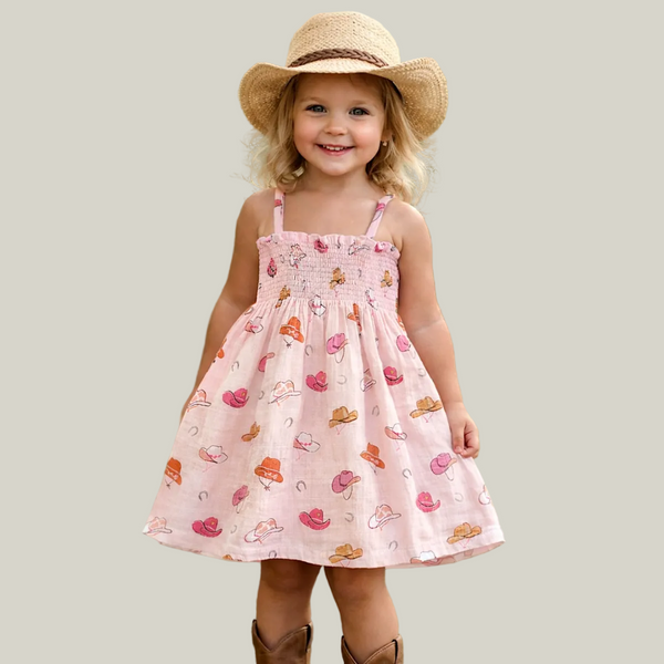 Child wearing a pink dress with cowboy hats and boots pattern, brown hat, and boots on a light gray background