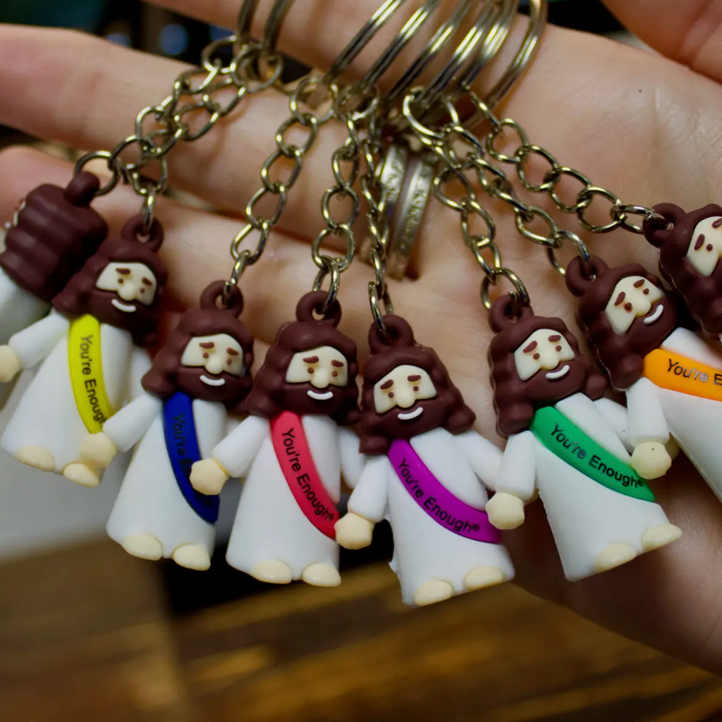A hand holding a set of small keychains shaped like Jesus in white robes with colored sashes and the words 'You Are Enough' on each one, attached to gold-tone rings.