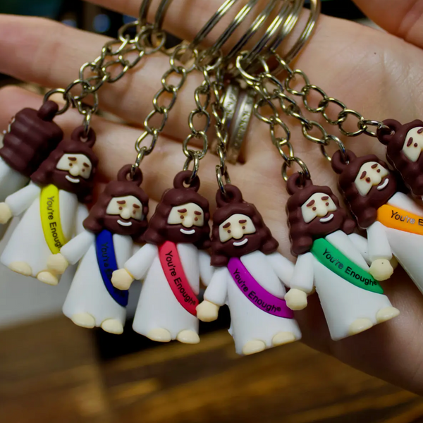 A hand holding a set of small keychains shaped like Jesus in white robes with colored sashes and the words 'You Are Enough' on each one, attached to gold-tone rings.