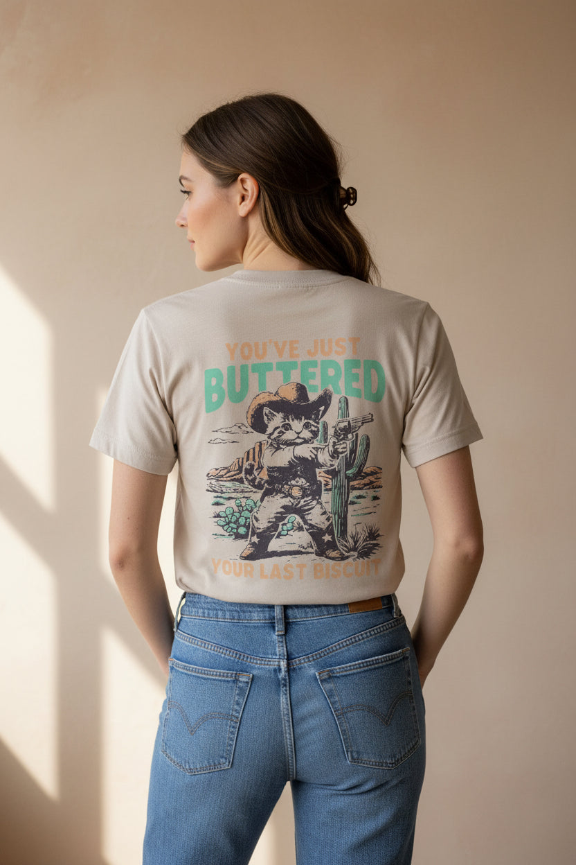 A beige graphic t-shirt hanging on a wooden hanger in a store. The back of the shirt features a western-themed illustration of a cat dressed as a cowboy holding a gun and a cactus, with the text 'YOU'VE JUST BUTTERED' and 'YOUR LAST BISCUIT'.