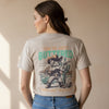 A beige graphic t-shirt hanging on a wooden hanger in a store. The back of the shirt features a western-themed illustration of a cat dressed as a cowboy holding a gun and a cactus, with the text 'YOU'VE JUST BUTTERED' and 'YOUR LAST BISCUIT'.