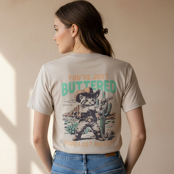 A beige graphic t-shirt hanging on a wooden hanger in a store. The back of the shirt features a western-themed illustration of a cat dressed as a cowboy holding a gun and a cactus, with the text 'YOU'VE JUST BUTTERED' and 'YOUR LAST BISCUIT'.