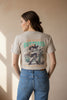 A beige graphic t-shirt hanging on a wooden hanger in a store. The back of the shirt features a western-themed illustration of a cat dressed as a cowboy holding a gun and a cactus, with the text 'YOU'VE JUST BUTTERED' and 'YOUR LAST BISCUIT'.