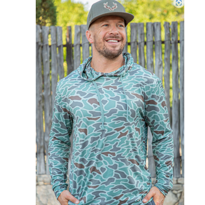 A man wearing a green camouflage performance hoodie with the hood up, standing in front of a wooden fence.